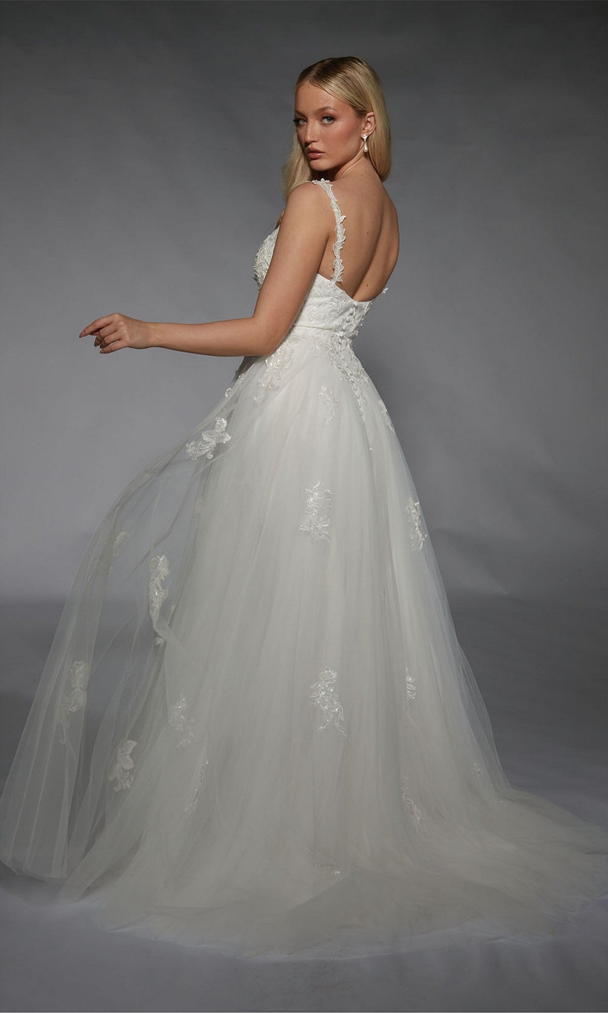 Woman wearing the Alyce 7124 long white dress with lace details, standing against a gray background and looking over her shoulder.